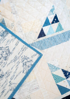 An upclose view of a patchwork quilt in a variation of blues and neutrals forming little sailboats. Quilt binding can be seen with hand done stitches.