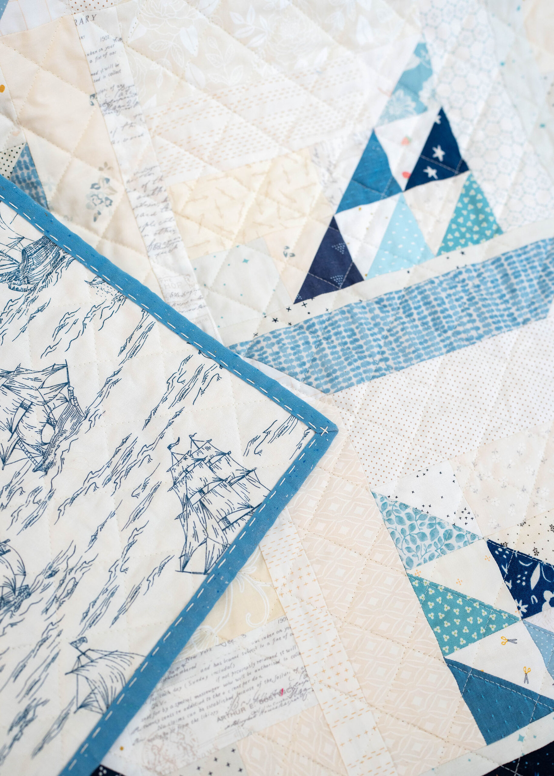 An upclose view of a patchwork quilt in a variation of blues and neutrals forming little sailboats. Quilt binding can be seen with hand done stitches.